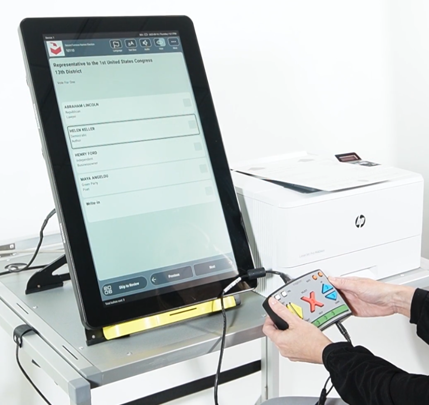 A person using an electronic voting machine with a touchscreen interface and a handheld controller. The screen displays a ballot for the 1st United States Congress, 13th District, with candidate names and a write-in option. A printer is visible in the background.